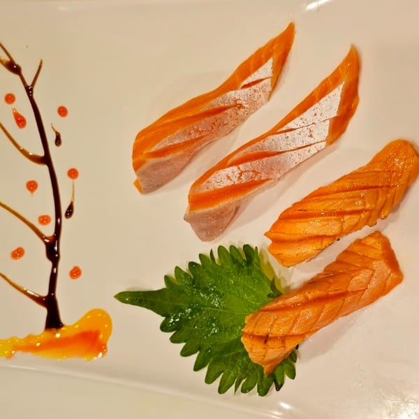 Four pieces of salmon sashimi on a white plate with a green leaf and sauce art.