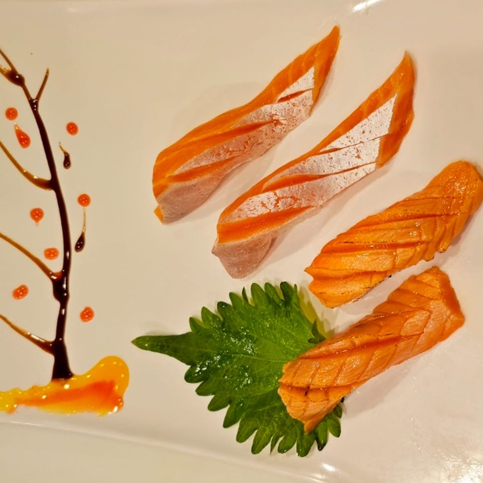 Four pieces of salmon sashimi on a white plate with a green leaf and sauce art.